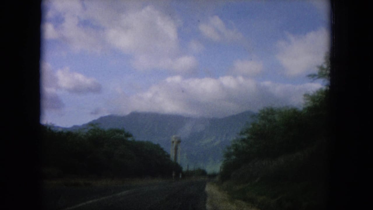 1964 Oahi, Hawaii road-view frame with a paved lane, green vegetation, distant ridges, clouds, and a small tower visible ahead.