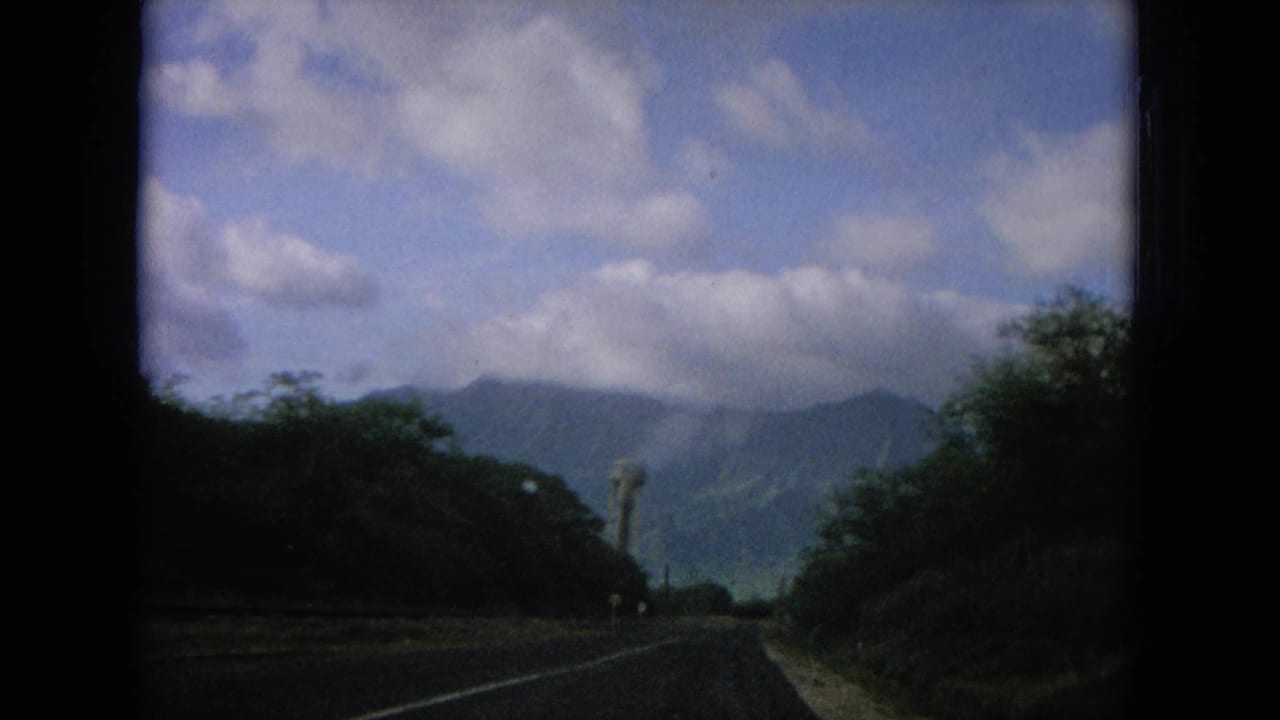 1964 Oahi, Hawaii driving footage frame showing a two-lane road between vegetation, distant mountains, clouds, and a tower at roadside.