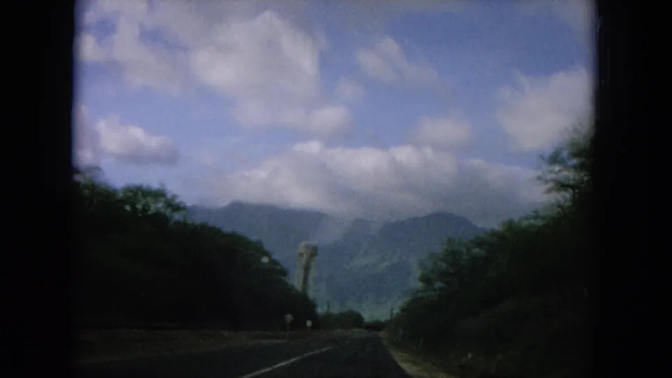 1964 Oahi, Hawaii home-movie frame looking down a paved road toward green mountain slopes, clouds, and a roadside tower.