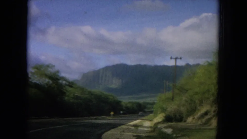 1964 Oahi, Hawaii frame showing a curving road, utility poles, lush roadside growth, a distant cliff face, and cloudy sky.