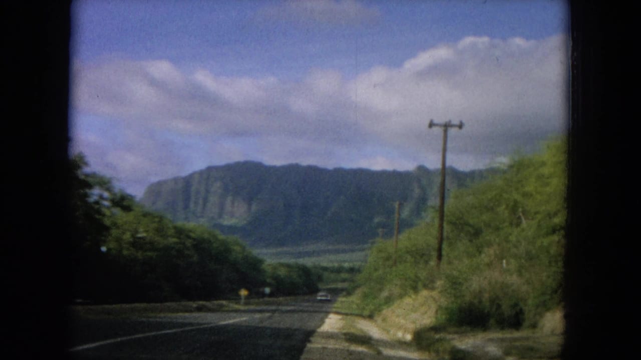 Paved road with green roadside vegetation, utility poles, distant mountains, blue sky, and white clouds.