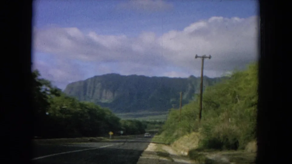 1964 Oahu home-movie frame showing a roadside view toward green mountain ridges beneath blue sky and scattered white clouds.