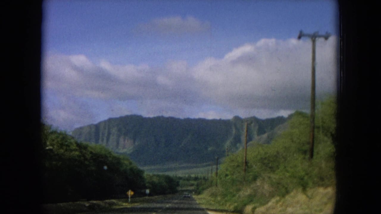 Roadside vegetation and utility poles frame distant green mountains under a clouded blue sky.