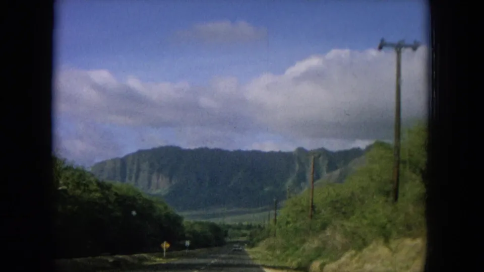 1964 Oahu travel footage frame with a road, utility poles, dense greenery, and distant ridges under bright clouds.