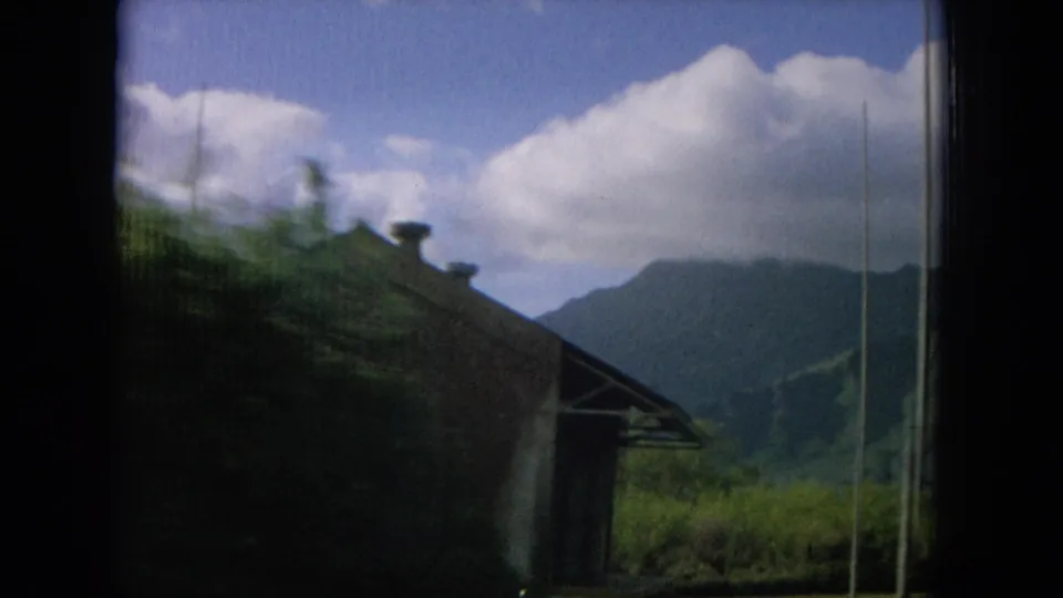 1964 Oahu home-movie frame showing a partial roadside building or shelter, tall grass, mountains, and large white clouds.