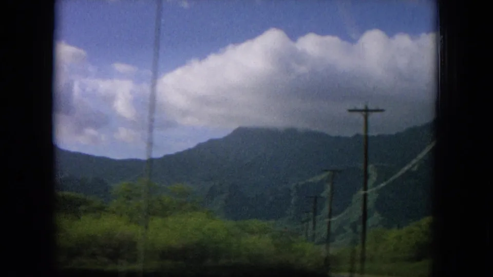 1964 Oahu travel frame looking past roadside poles and green vegetation toward a shaded mountain ridge under clouds.