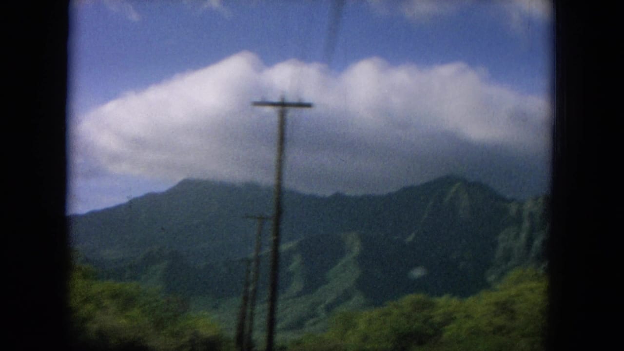 Green mountain ridge seen past roadside utility poles under a cloudy blue sky.