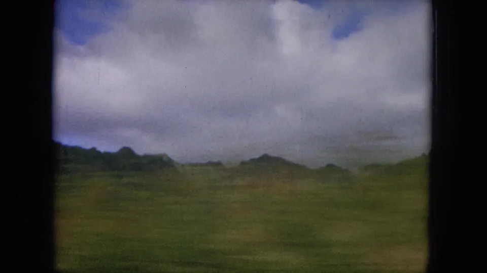 1964 Hawaiian landscape from a moving vehicle, showing open grassland, distant ridges, and a cloudy sky.