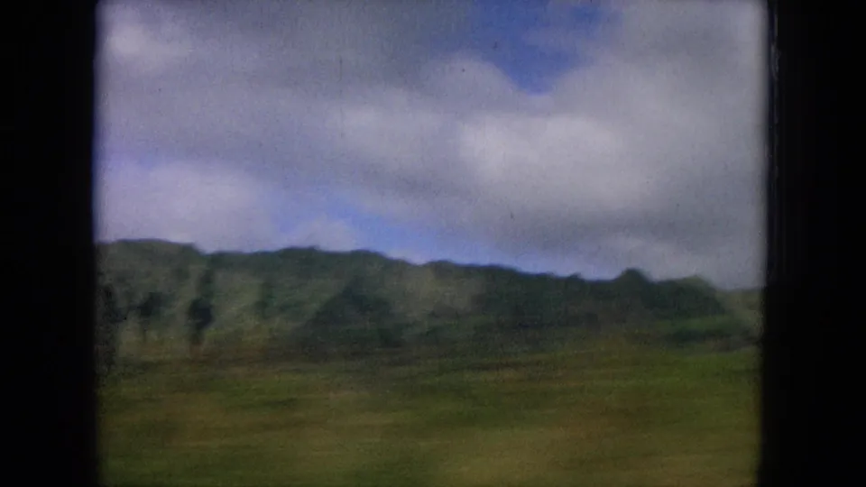 Vintage 8mm view of open Hawaiian terrain with distant green mountains and broad clouds overhead.