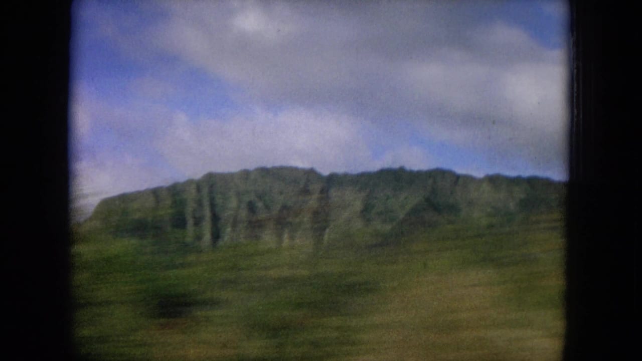 Green mountain ridge seen through dark vehicle-window framing under a blue, cloudy sky.