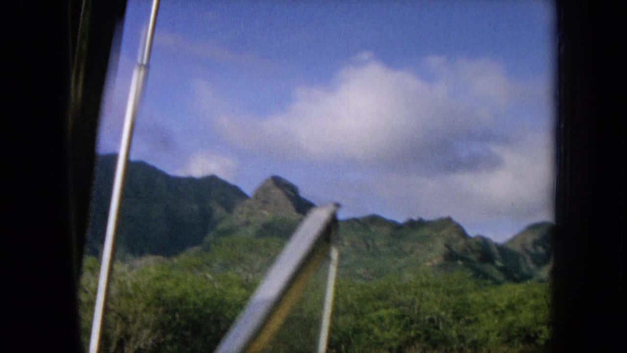 Tropical mountains and brush seen past a slanted metal rail or windshield edge.