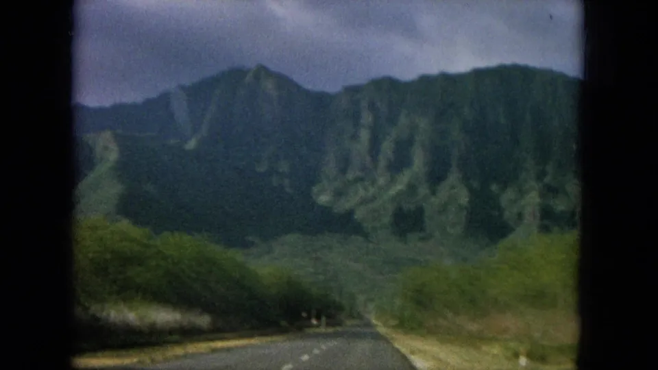 1960s 8mm Hawaii driving view along a paved road toward steep green mountains and roadside foliage.