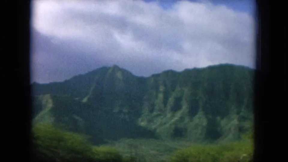 Vintage Hawaii home-movie frame of green mountain slopes and tropical vegetation under scattered clouds.