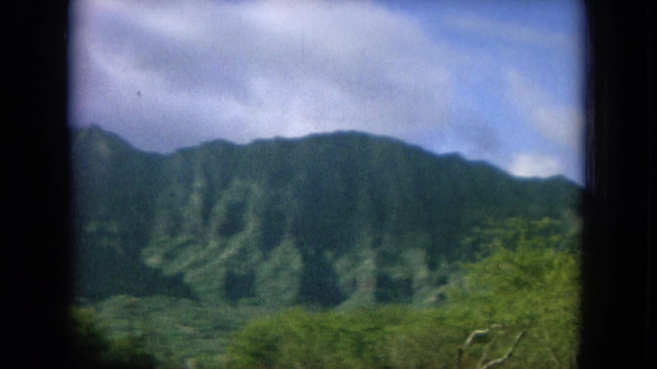 1964 8mm Hawaii travel frame showing green Oahu mountains through a dark window frame, with shrubs below and clouds above.