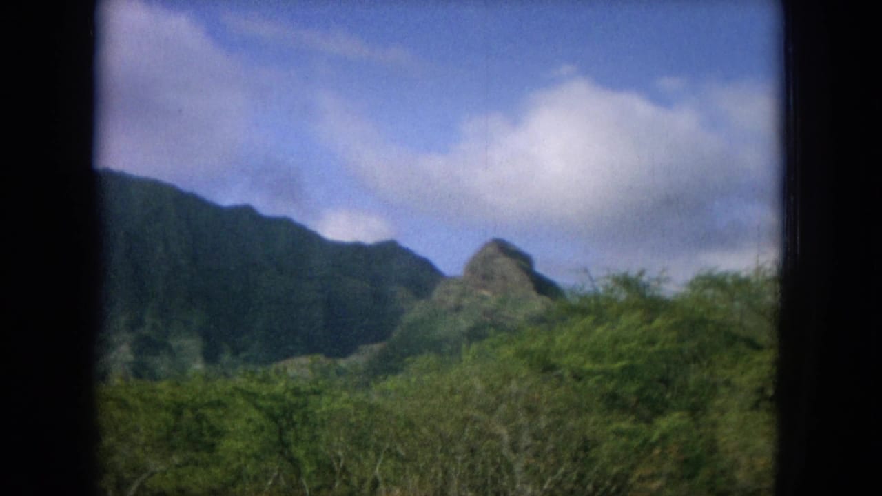 1964 Oahu home-movie frame with lush mountain ridges, a small pointed peak, foreground shrubs, and dark window borders.