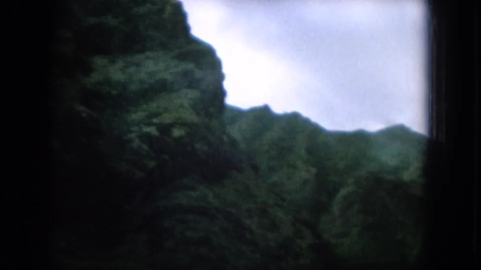 1964 8mm Oahu travel frame showing a shadowed green mountainside, distant ridge, dark window edge, and bright overcast sky.