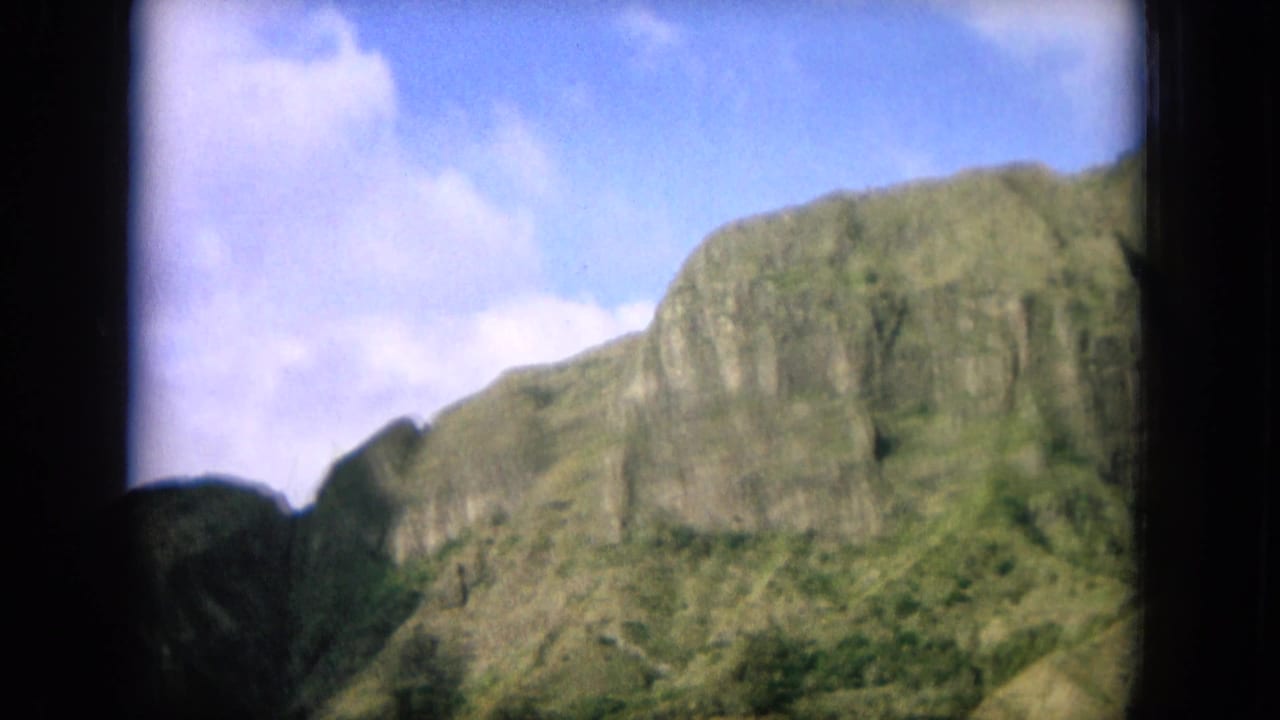 1964 Oahu home-movie frame showing a green mountain cliff through a dark window border beneath scattered clouds.