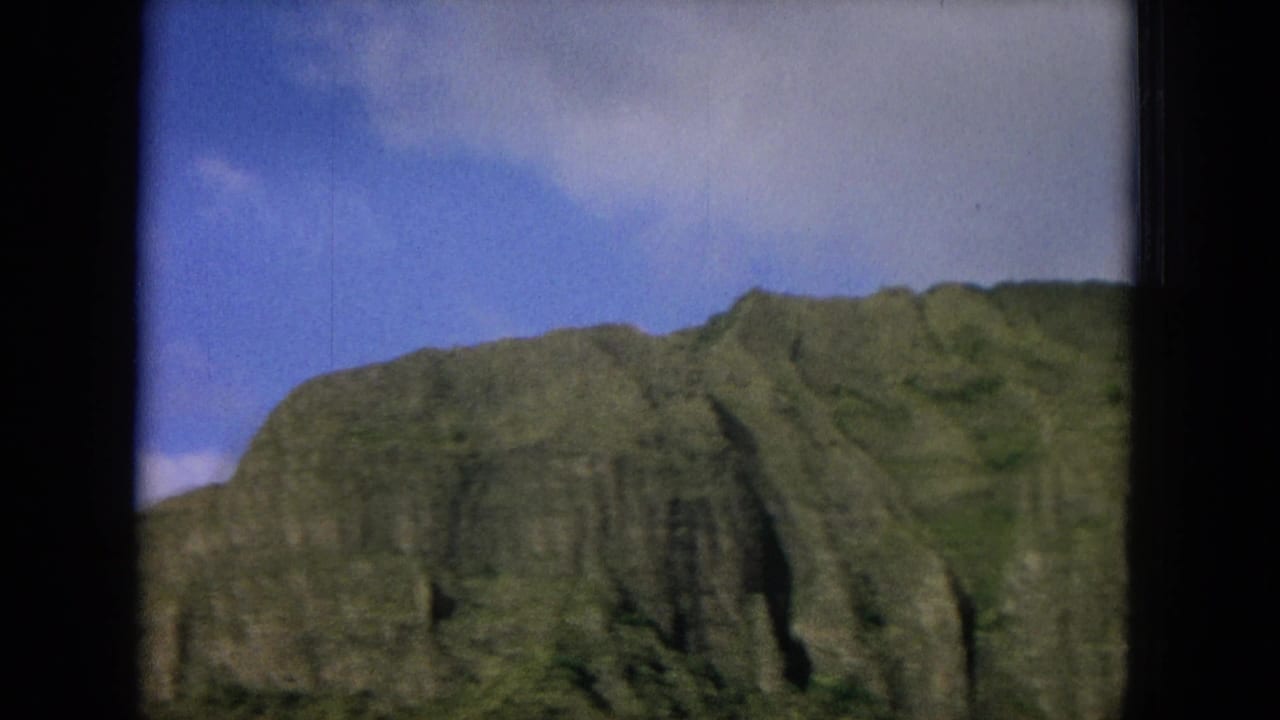 1964 Oahu travel footage frame with a rugged green cliff face framed by dark window edges and bright sky.