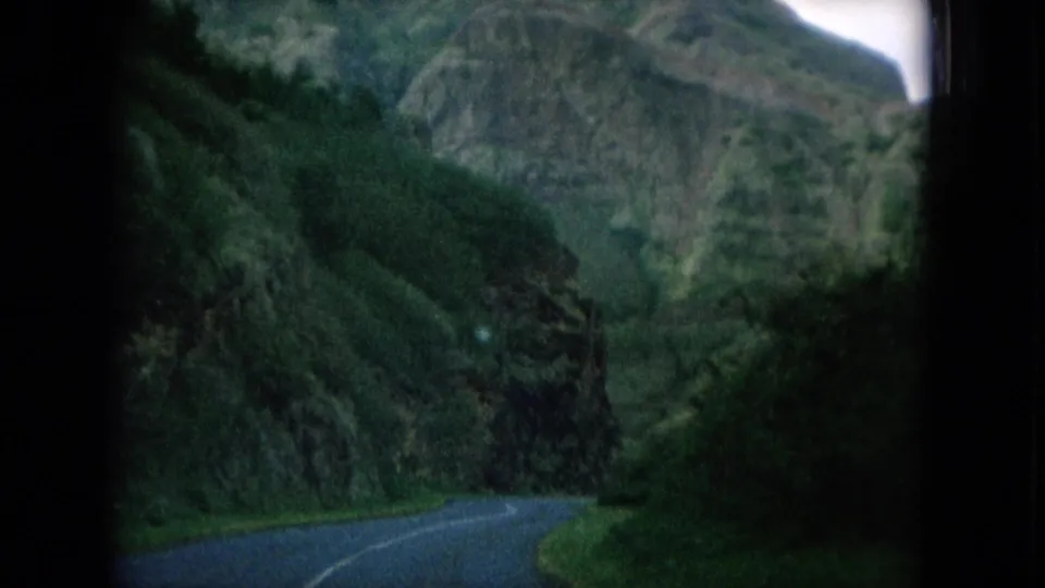 1964 Oahu home-movie frame showing a curving road and steep green mountains from a moving visitor viewpoint.