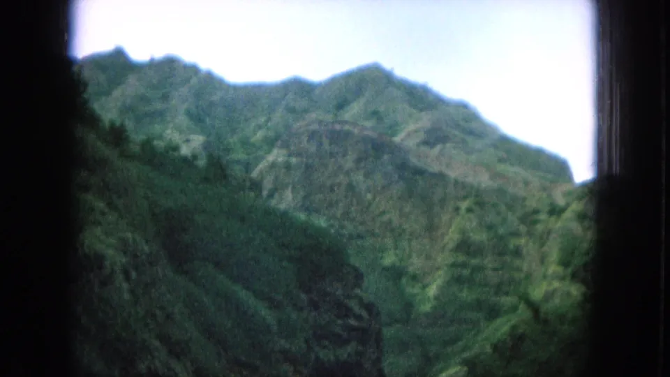 1964 Oahu frame with layered green ridges, rugged cliffs, and bright sky seen through a dark window border.