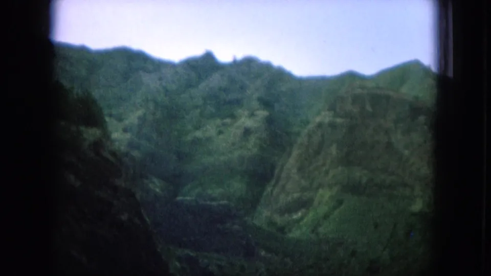 1964 8mm Hawaii travel frame showing lush mountain ridges through a dark window frame with soft focus and muted color.