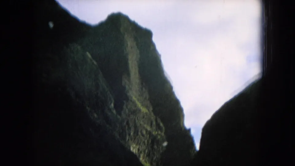1964 Hawaii home-movie frame with a steep green mountain face partly silhouetted between dark window borders.