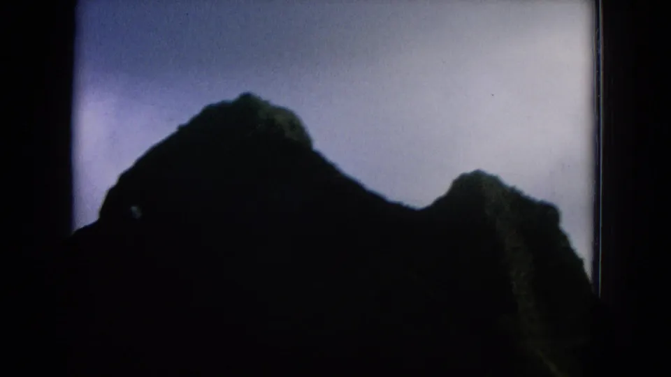 1964 8mm Hawaii frame showing dark mountain silhouettes and green ridges beneath a pale blue-gray sky.