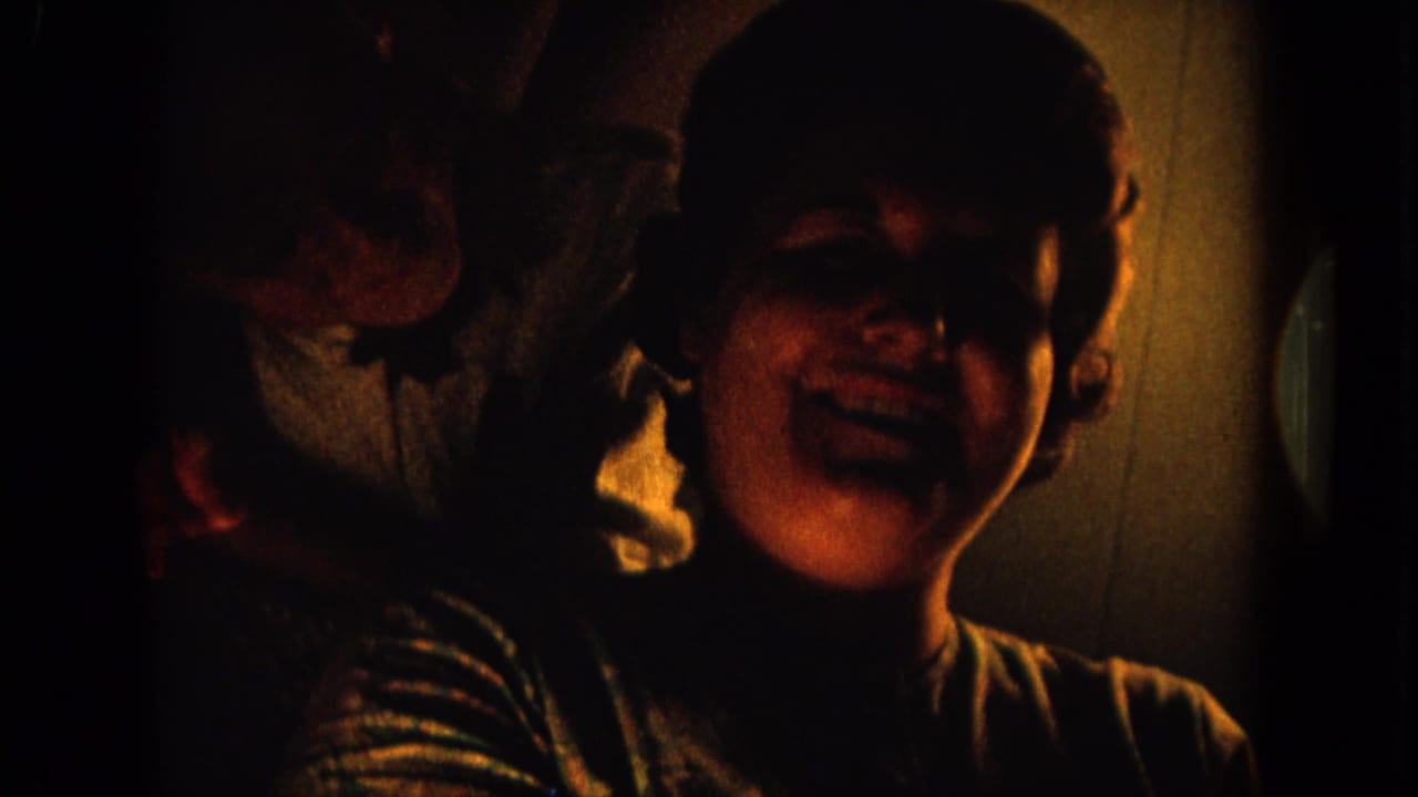 1964 8mm home-movie frame from Hawaii showing an adult woman smiling in low interior light, with a partial person at the left edge.