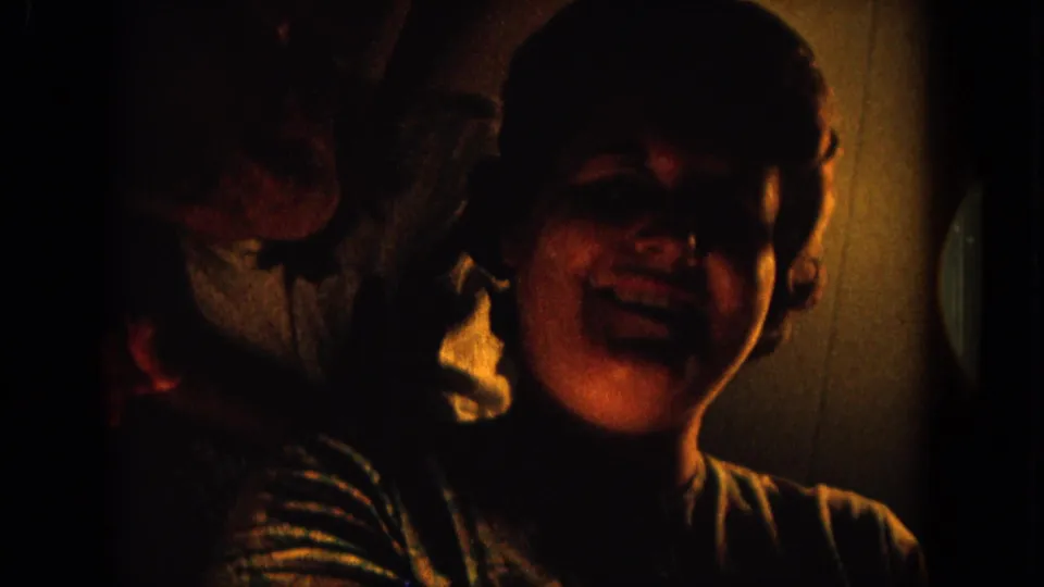 1964 8mm home-movie frame from Hawaii showing an adult woman smiling in low interior light, with a partial person at the left edge.