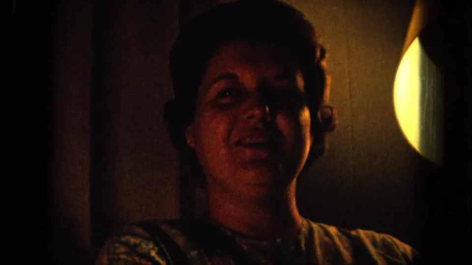 1964 8mm home-movie frame from Hawaii showing an adult woman facing the camera in warm low light beside a circular glow.