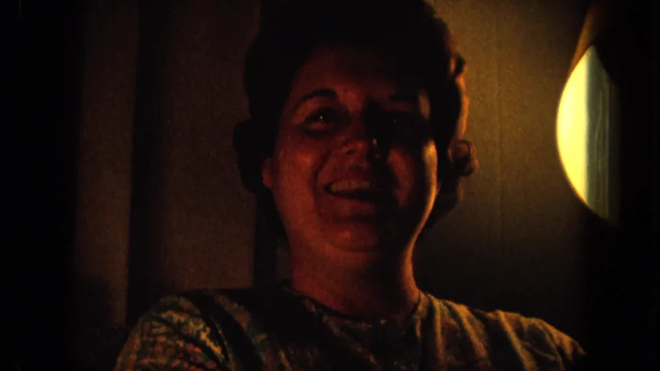 1964 Hawaii 8mm frame of an adult woman smiling in a dark private room with a bright circular light on the right.