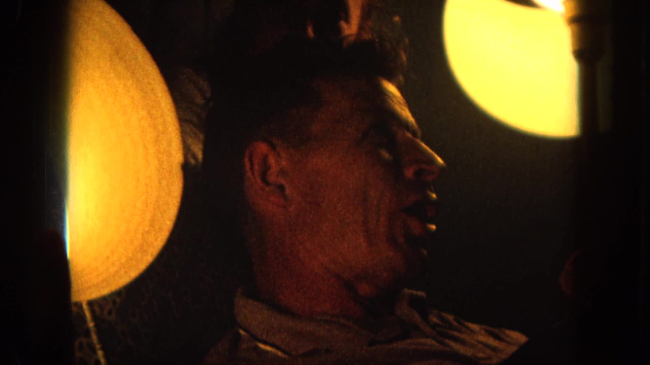 1964 8mm Hawaii home-movie frame of a man in profile speaking in warm low light beside circular lamps.