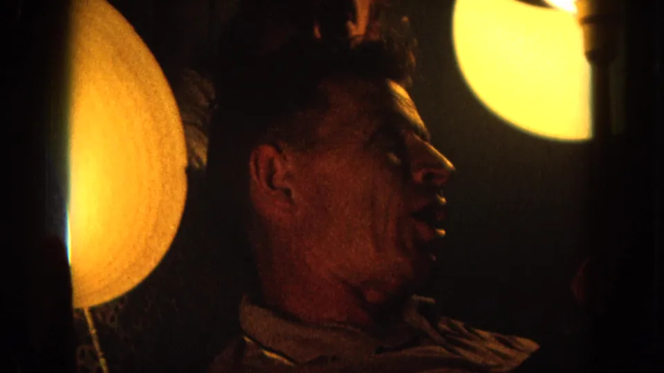 1964 8mm Hawaii home-movie frame of a man in profile speaking in warm low light beside circular lamps.