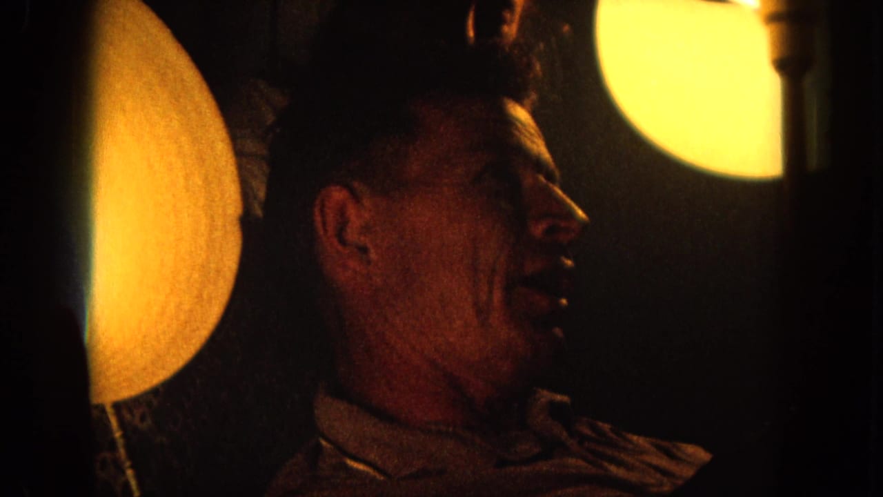 Warm 1964 8mm frame showing the man in side profile, seated or reclining in a dark private interior.