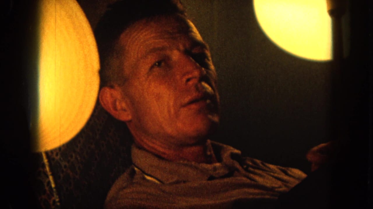 1964 Hawaii 8mm frame of a seated adult man looking upward beside a glowing round lamp in a dim interior.