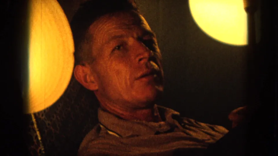 1964 Hawaii 8mm frame of a seated adult man looking upward beside a glowing round lamp in a dim interior.