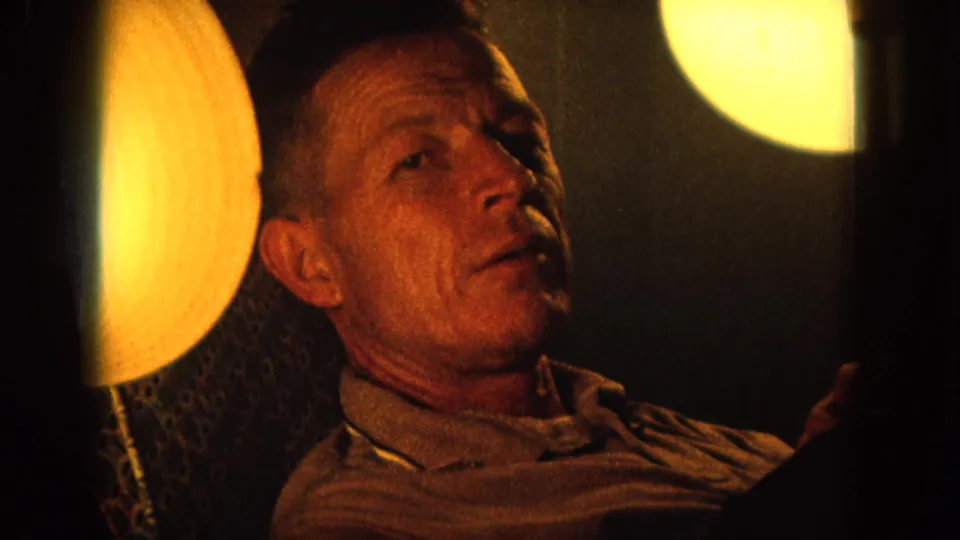 Close 1964 Hawaii 8mm view of a seated man in a collared shirt beside a warm circular lamp and dark wall.