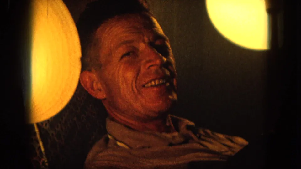 1964 Hawaii 8mm frame of a seated man faintly smiling in a dim private room with warm lamp light.