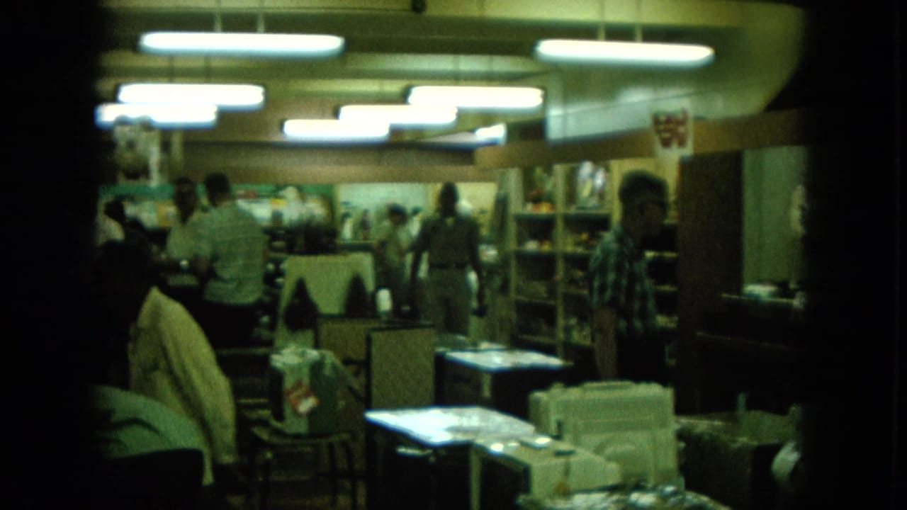1960s Hawaii home-movie frame showing a dim store interior with people browsing beneath bright fluorescent lights.