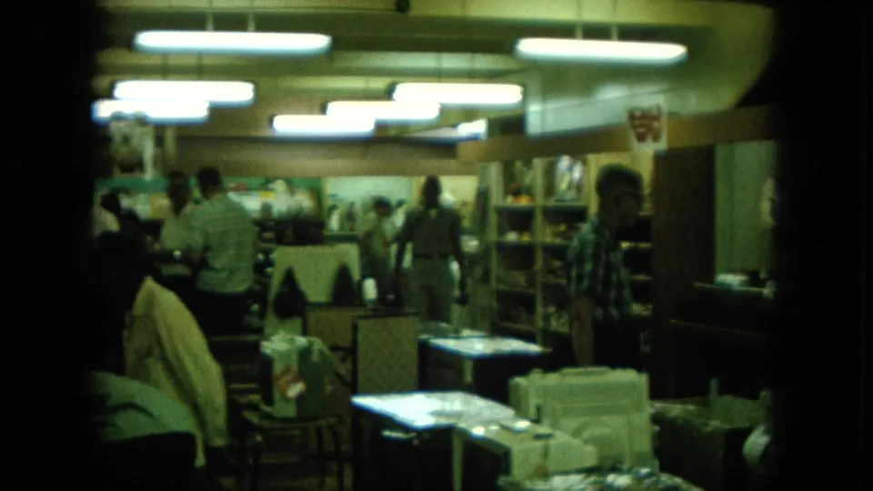 1960s Hawaii home-movie frame showing a dim store interior with people browsing beneath bright fluorescent lights.