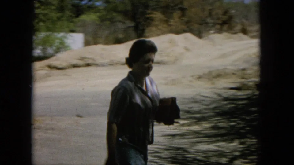 Woman in a light top walks along a shoreline or rocky water edge in Hawaii, carrying a purse in handheld 8mm footage.
