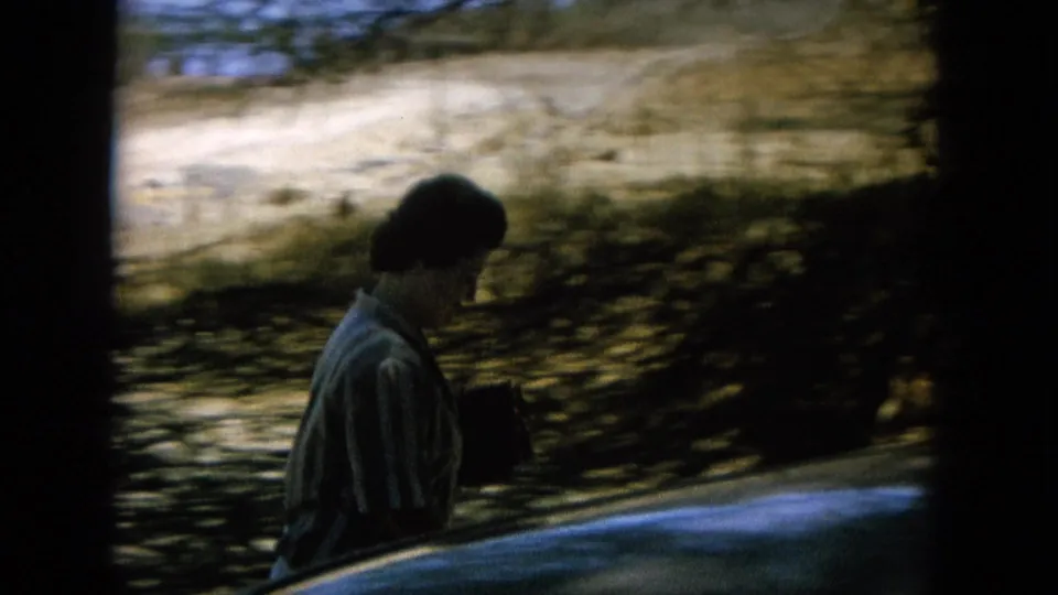 Home-movie frame of a woman walking near a shaded shoreline, partly framed by a car edge in the foreground.