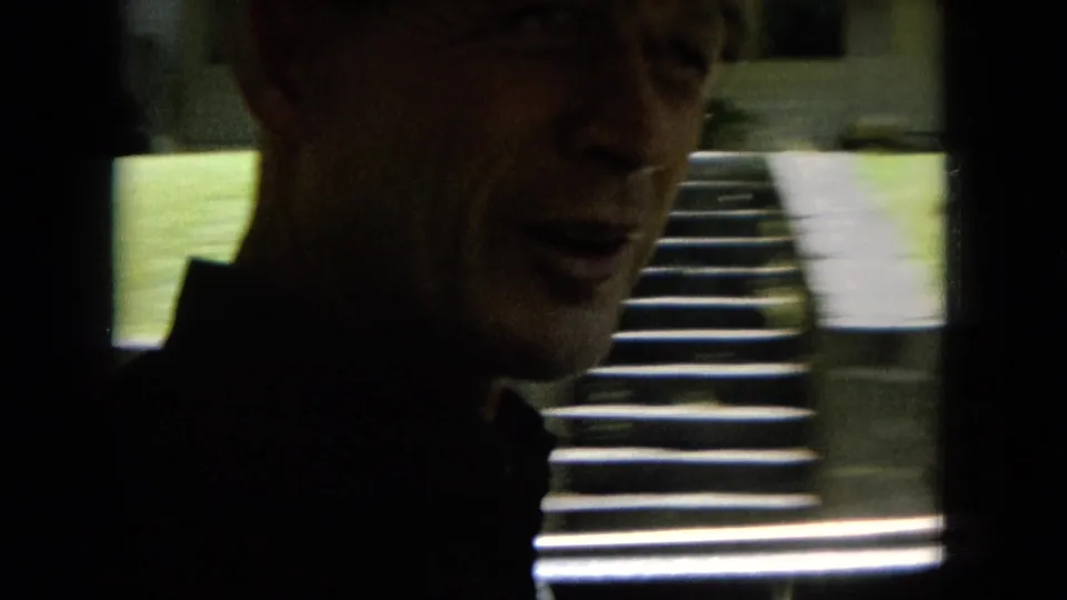 Close handheld 8mm view of a man smiling near a vehicle window or opening, with outdoor steps visible in the background.