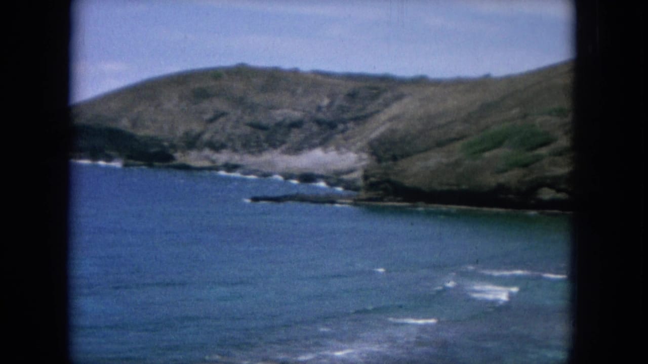 Rocky shoreline and green-brown hills above blue water with small white surf lines.