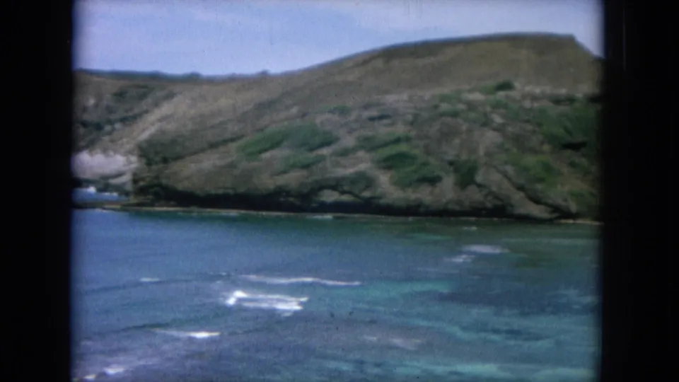Oahu coastal cove area on 8mm film, with turquoise shallows, reef-like patches, surf streaks, and steep hills.