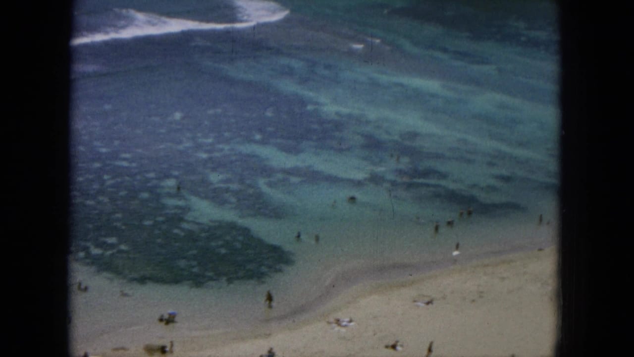 1964 8mm-style elevated view of an Oahu beach, with small swimmers in shallow blue-green water and people on the sand.