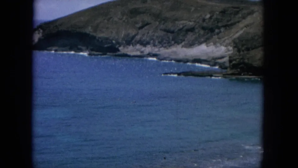 1964 8mm coastal view in Hawaii with a rugged hillside, rocky shoreline, and calm blue ocean framed by the film gate.