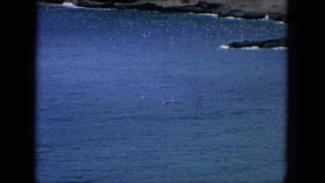 1964 Oahu home-movie frame showing open blue ocean and a dark rocky shoreline edge within visible 8mm gate borders.