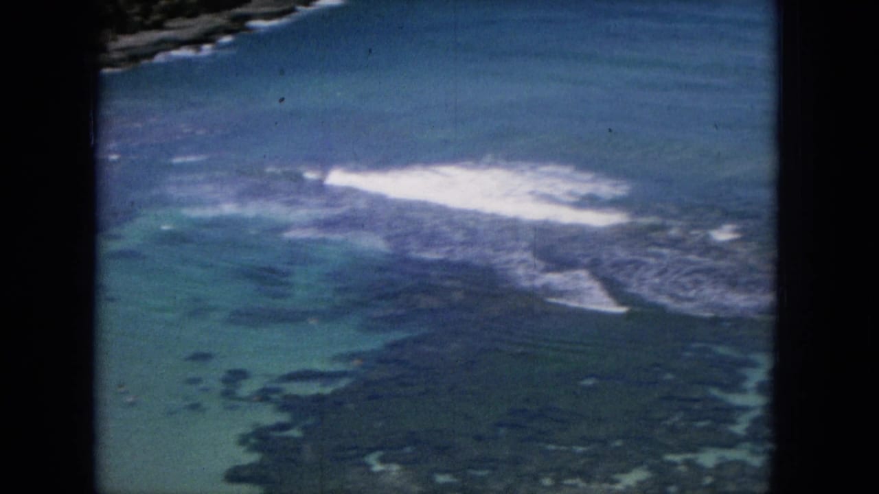 1964 Oahu coastal view with turquoise shallows, dark reef or rock patches, and white surf breaking near the shoreline.