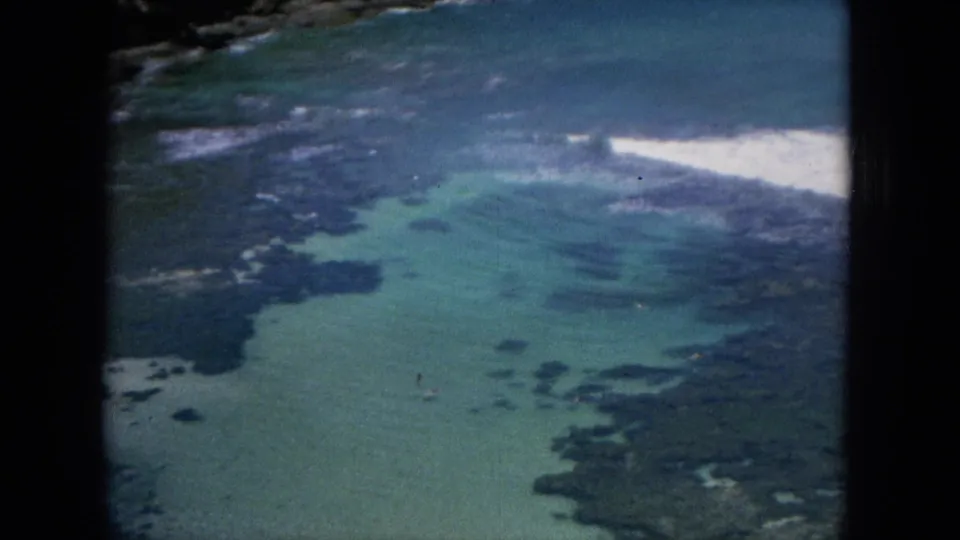 1964 Oahu shoreline frame showing shallow turquoise water, submerged dark formations, and surf breaking beside the coast.